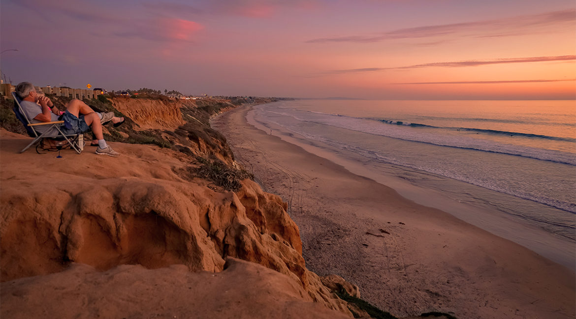 Stunning sunset view from a cliff overlooking a Southern California beach, showcasing the coastal lifestyle of Ventura and Los Angeles Counties, perfect for real estate in 2025 buyer’s market, with a person relaxing.