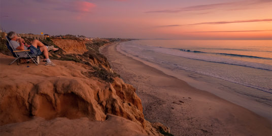 Stunning sunset view from a cliff overlooking a Southern California beach, showcasing the coastal lifestyle of Ventura and Los Angeles Counties, perfect for real estate in 2025 buyer’s market, with a person relaxing.