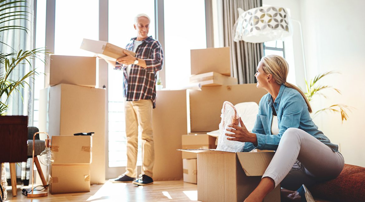 Two people in a cozy living room packing labeled boxes with books, photos, and clothes, illustrating downsizing preparation for a 2025 home sale.
