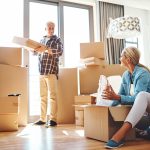 Two people in a cozy living room packing labeled boxes with books, photos, and clothes, illustrating downsizing preparation for a 2025 home sale.